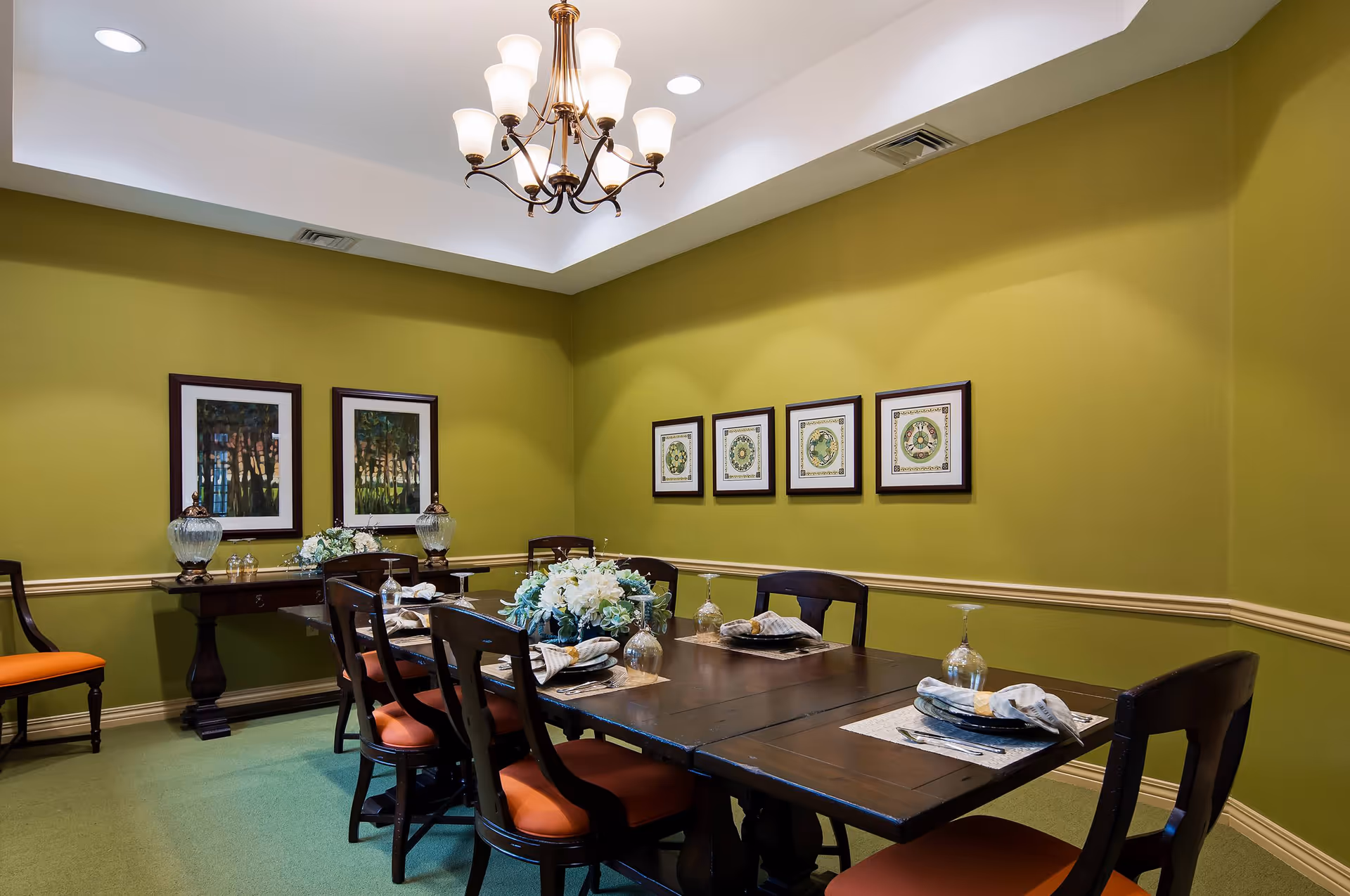 Formal dining room with a long wooden table set for meals, orange-cushioned chairs, green walls and framed artwork.
