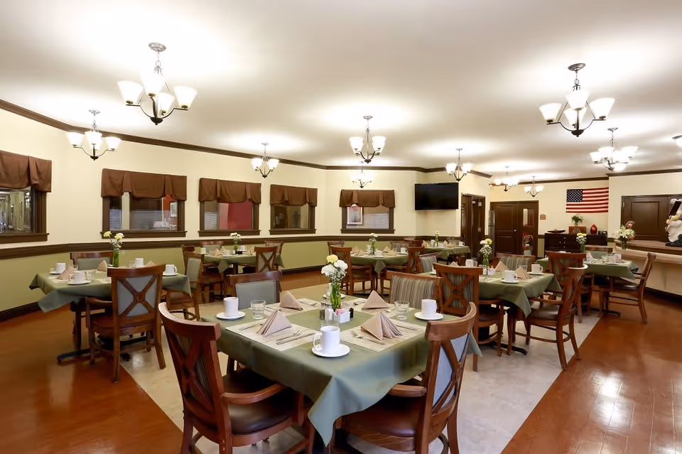 A well-lit dining room with multiple tables covered in green tablecloths, each set with white cups, glasses, napkins, and small flower vases. The room has wooden chairs, several windows with brown valances, chandeliers hanging from the ceiling, and an American flag on the far wall.