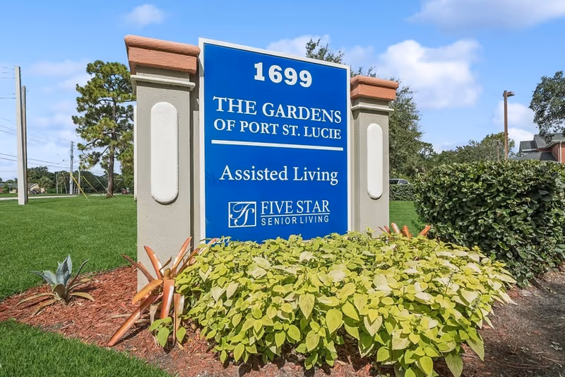 Outdoor sign for The Gardens of Port St. Lucie Assisted Living by Five Star Senior Living, surrounded by green plants and landscaping with a clear blue sky in the background.