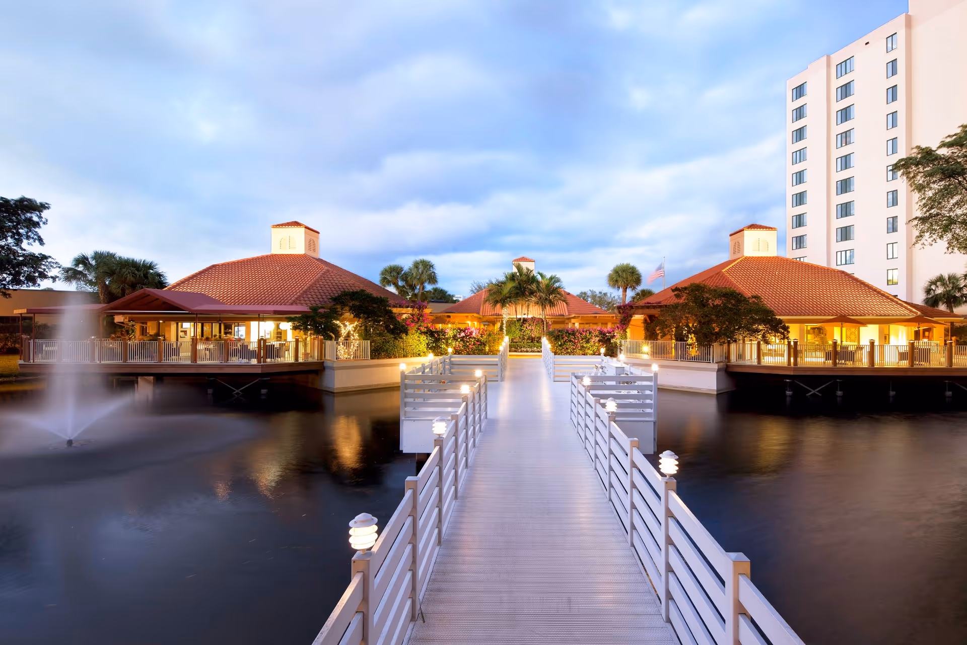 A scenic view of The Waterford facility featuring a long white wooden walkway with railings leading over a pond with a water fountain. On either side of the walkway are buildings with red-tiled roofs and outdoor seating areas surrounded by lush greenery and palm trees. A tall white building is visible in the background under a cloudy sky at dusk.