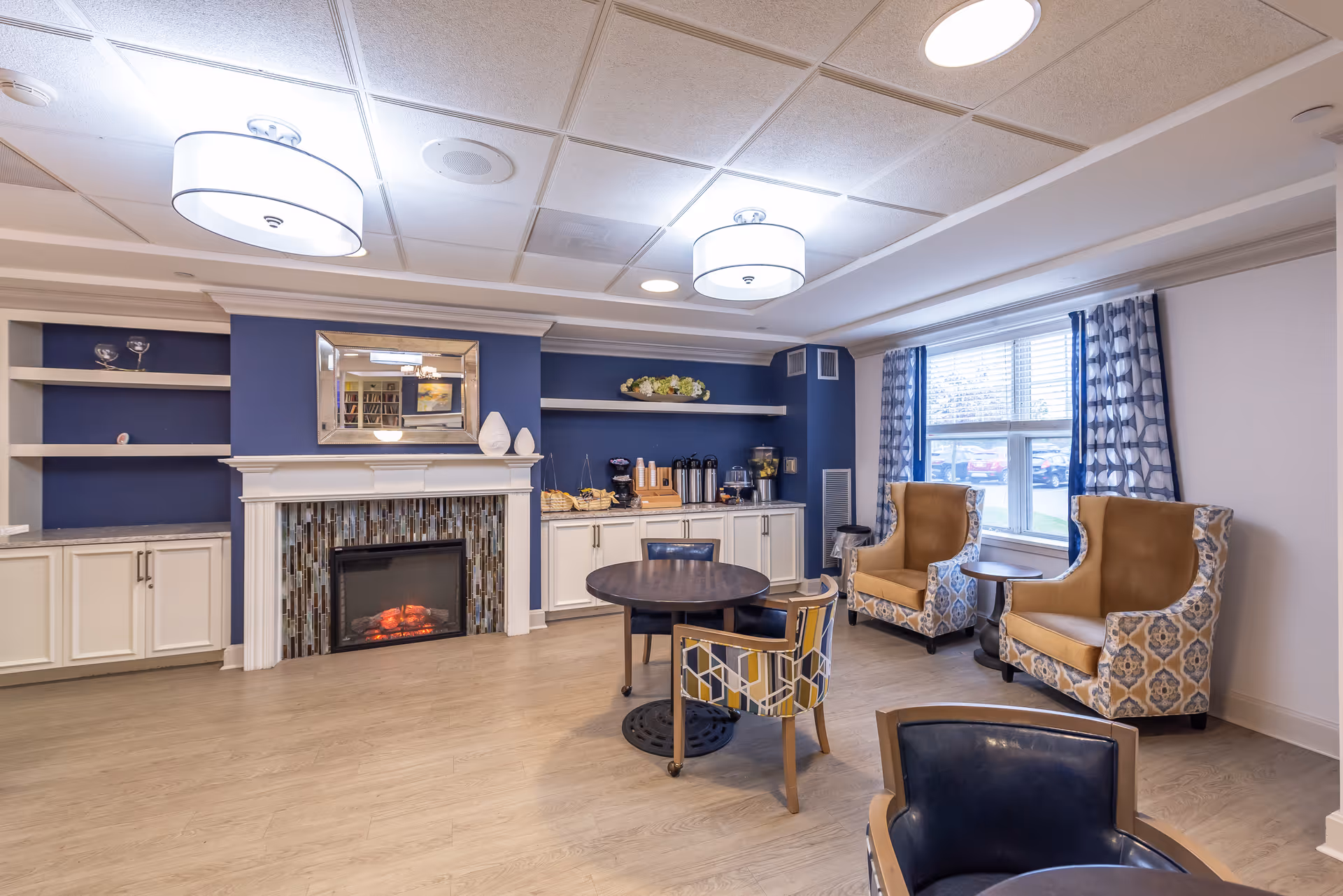 A cozy senior living common area with a modern electric fireplace set in a white mantel with mosaic tile surround. The wall behind the fireplace is painted dark blue with white shelves and cabinets on either side. There is a round wooden table with four chairs in the center of the room, and two patterned armchairs with mustard yellow cushions near a large window with blue and white patterned curtains. The room has light wood flooring and two large ceiling lights.