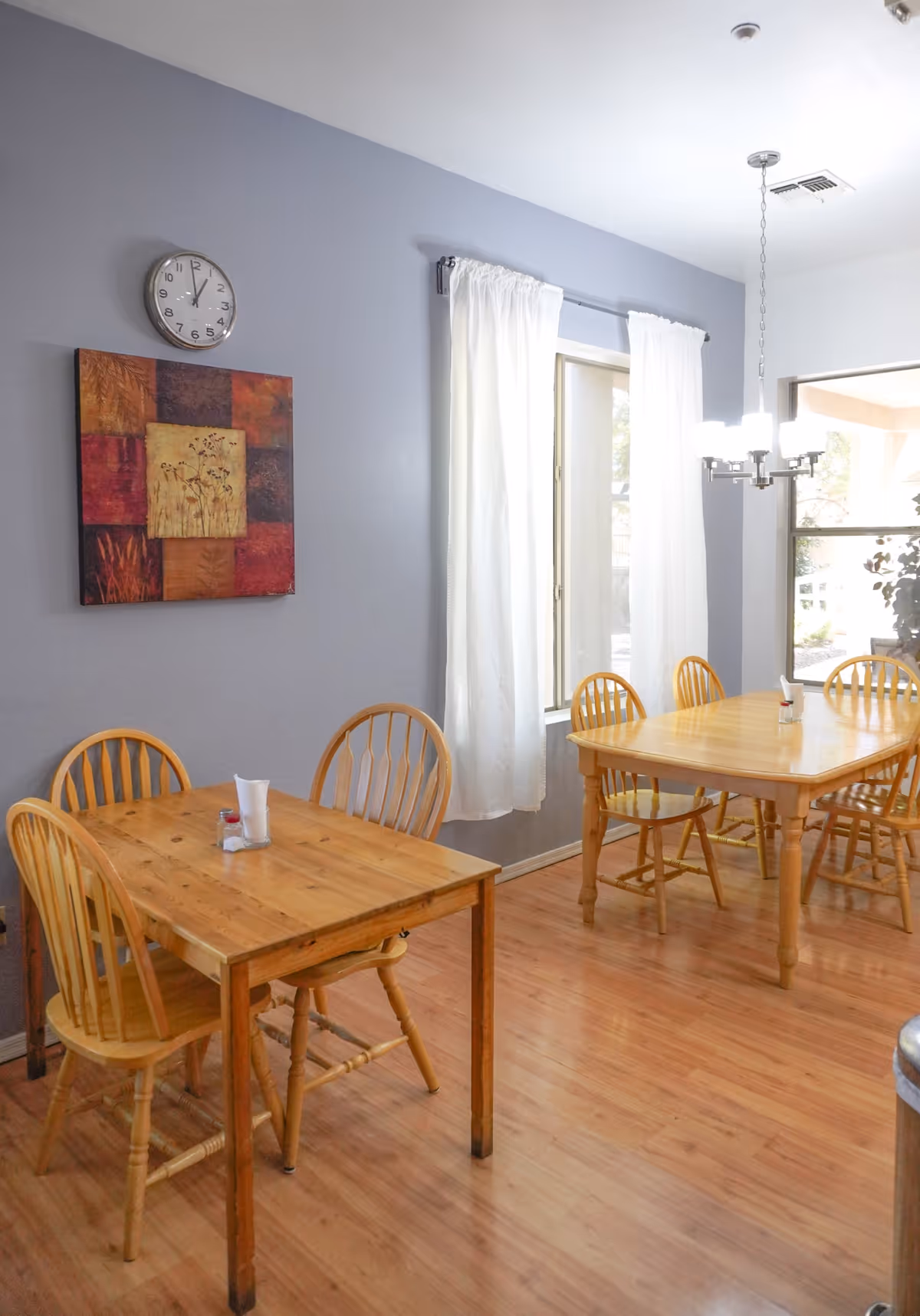 A dining area with two wooden tables and several wooden chairs on a light wood floor. The walls are painted gray with a colorful abstract painting and a clock hanging above one table. White curtains cover the windows, and a modern chandelier hangs above the larger table.