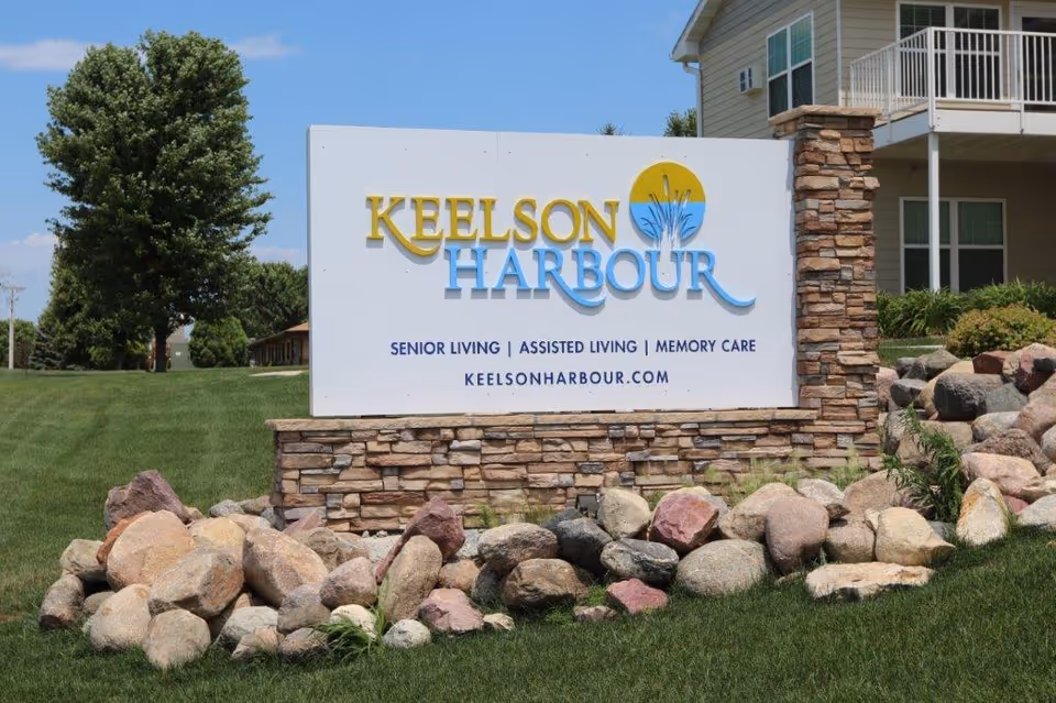 Outdoor view of a large sign for Keelson Harbour senior living facility, featuring the text 'Senior Living | Assisted Living | Memory Care' and the website keelsonharbour.com, surrounded by rocks and grass with a building and trees in the background under a clear blue sky.