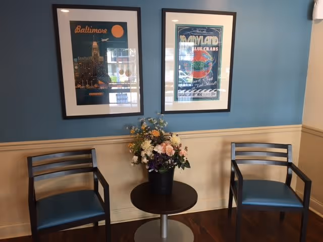 A small seating area with two wooden chairs with blue cushions on either side of a round table holding a flower arrangement. The wall behind is painted blue on the upper half and beige on the lower half, with two framed posters hanging above. One poster features Baltimore and the other features Maryland blue crabs.