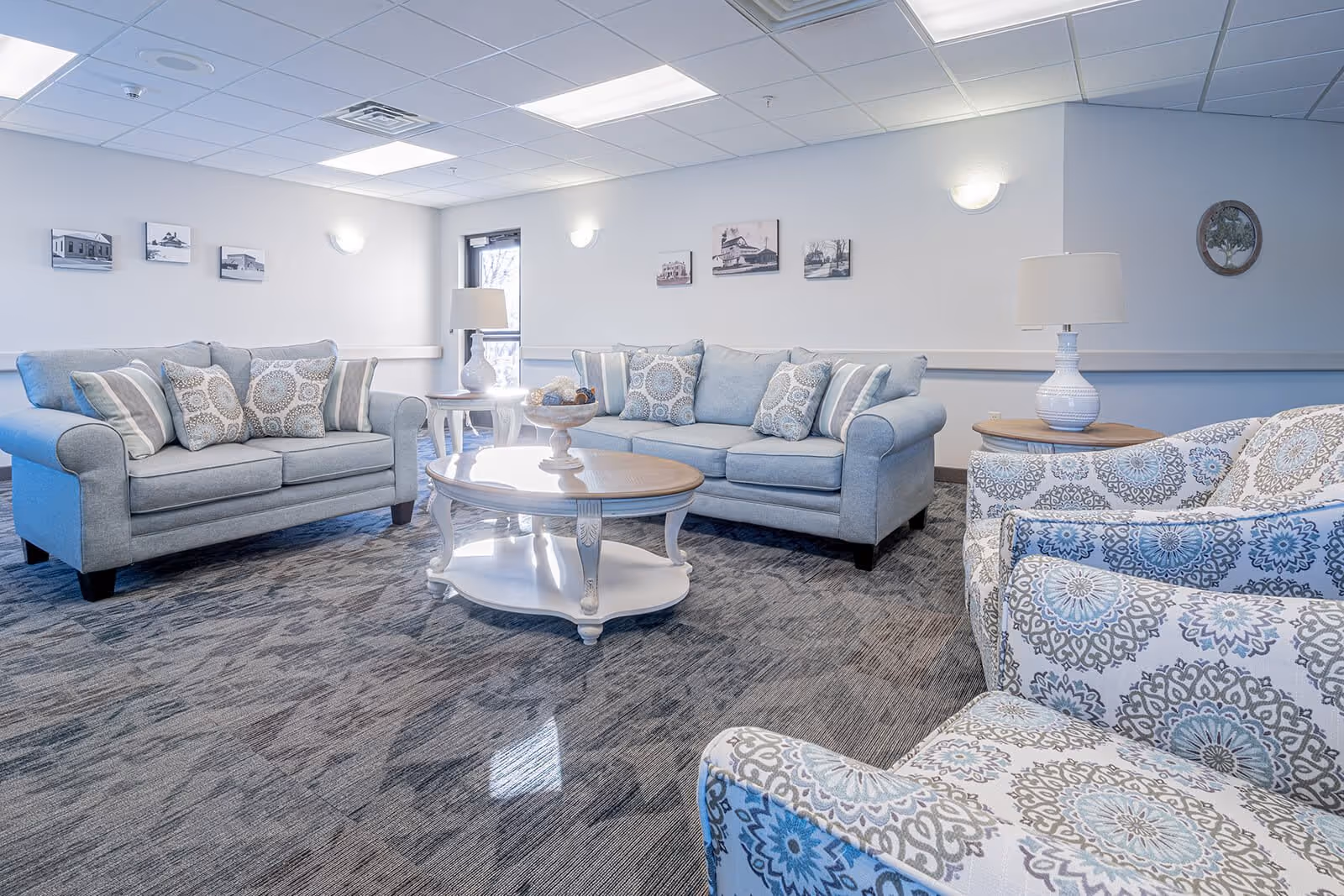 A bright and cozy living room area with two light blue sofas adorned with patterned throw pillows, two matching patterned armchairs, a white wooden coffee table with decorative items, and two white table lamps on side tables. The walls are decorated with framed pictures and a round wall art piece, and the room has a carpeted floor and a drop ceiling with fluorescent lighting.