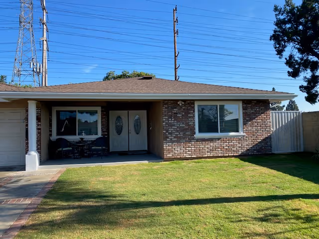 Single-story brick house front with a lawn, covered porch, double front doors, and power lines in the background.