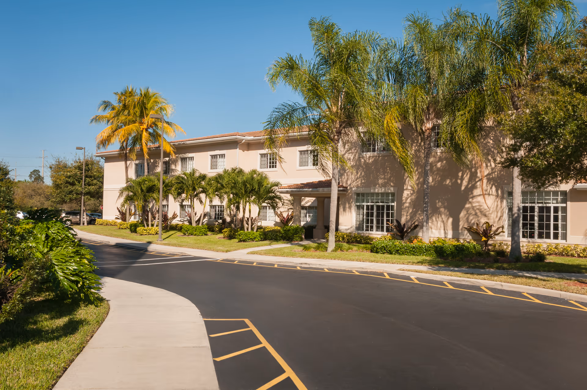 Two-story beige senior living building with palm trees, landscaping and a curved driveway under a clear blue sky.