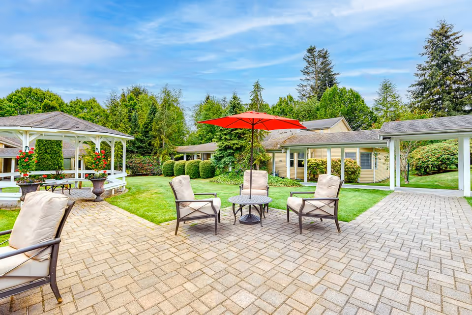 Outdoor patio area with four cushioned chairs arranged around a small round table with a red umbrella. The patio is paved with bricks and surrounded by green grass, bushes, and trees. A white gazebo with hanging flower pots is visible on the left side, and single-story buildings with beige siding and gray roofs are in the background under a partly cloudy sky.