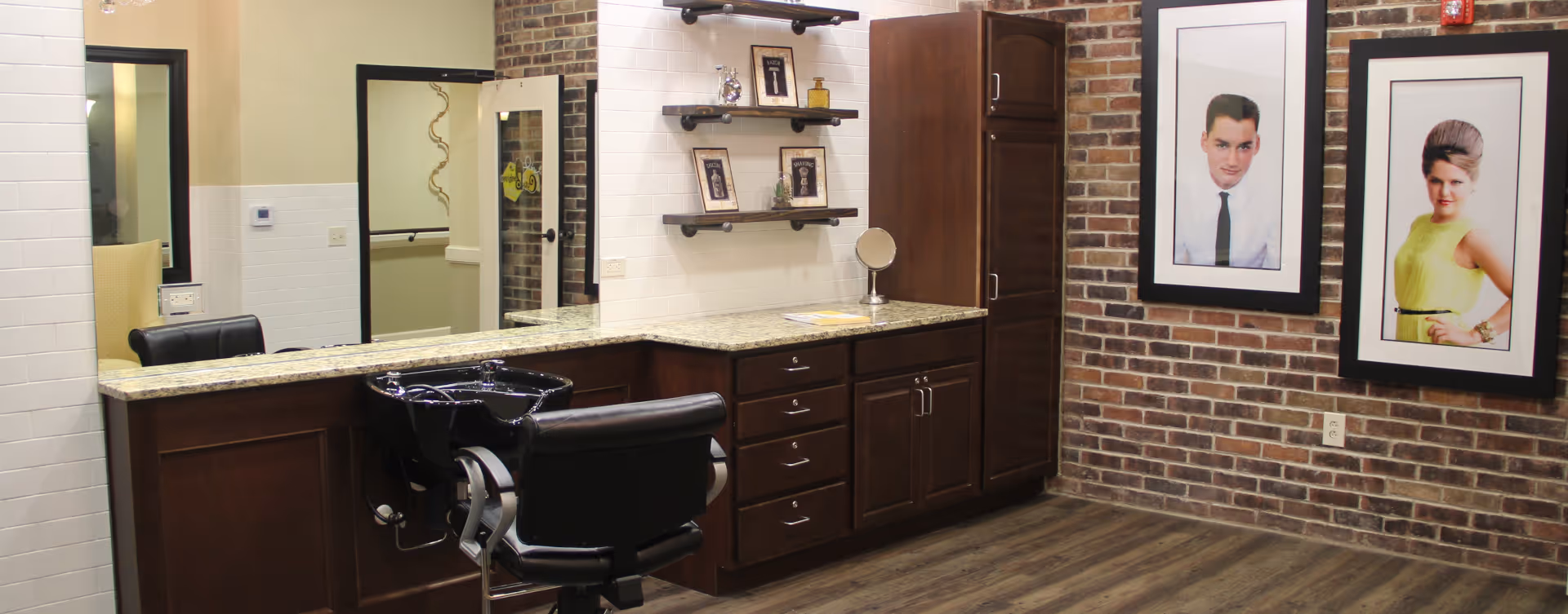 Interior view of a salon area with a black salon chair and a black hair washing sink attached to a dark wood counter with granite countertop. The wall behind the counter has white tiles and floating shelves with framed pictures and hair products. The adjacent wall is exposed brick with two large framed portraits of a man and a woman. The floor is wood, and there is a mirror reflecting part of the room and hallway.