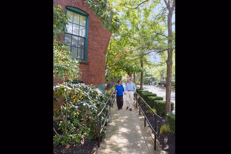 An elderly couple walks along a tree-lined sidewalk beside a brick building using handrails.