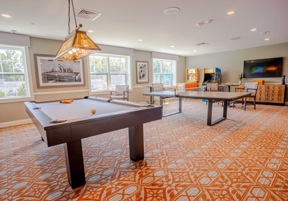 A bright recreational room with a pool table, a ping pong table, two arcade game machines, several chairs, a large flat-screen TV mounted on the wall, and large windows letting in natural light. The room has patterned carpet and framed black and white photos on the walls.