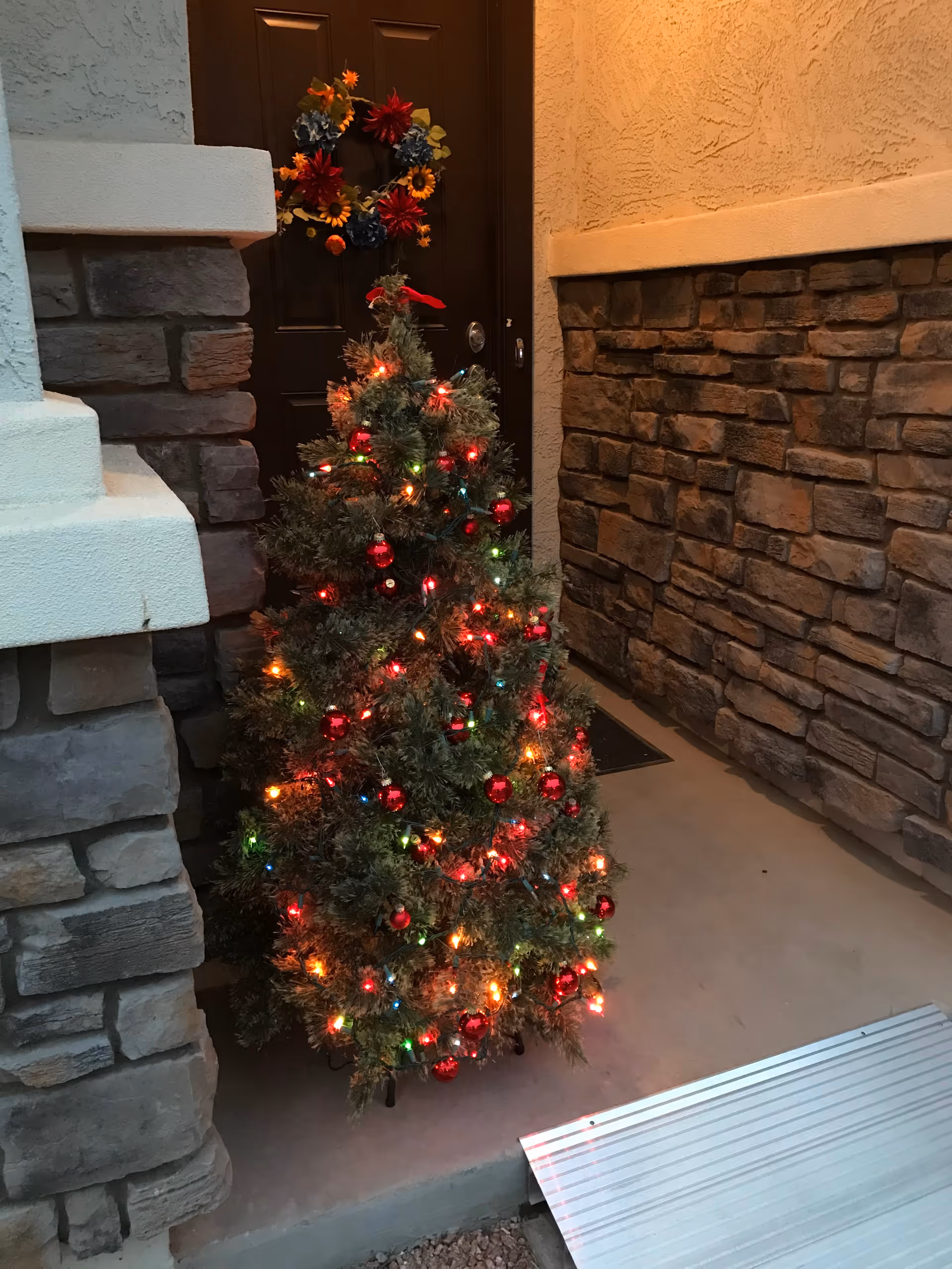 A small decorated Christmas tree with colorful lights and red ornaments placed outside near a dark brown door with a fall-themed wreath. The area is surrounded by stone and stucco walls with a metal ramp leading up to the entrance.