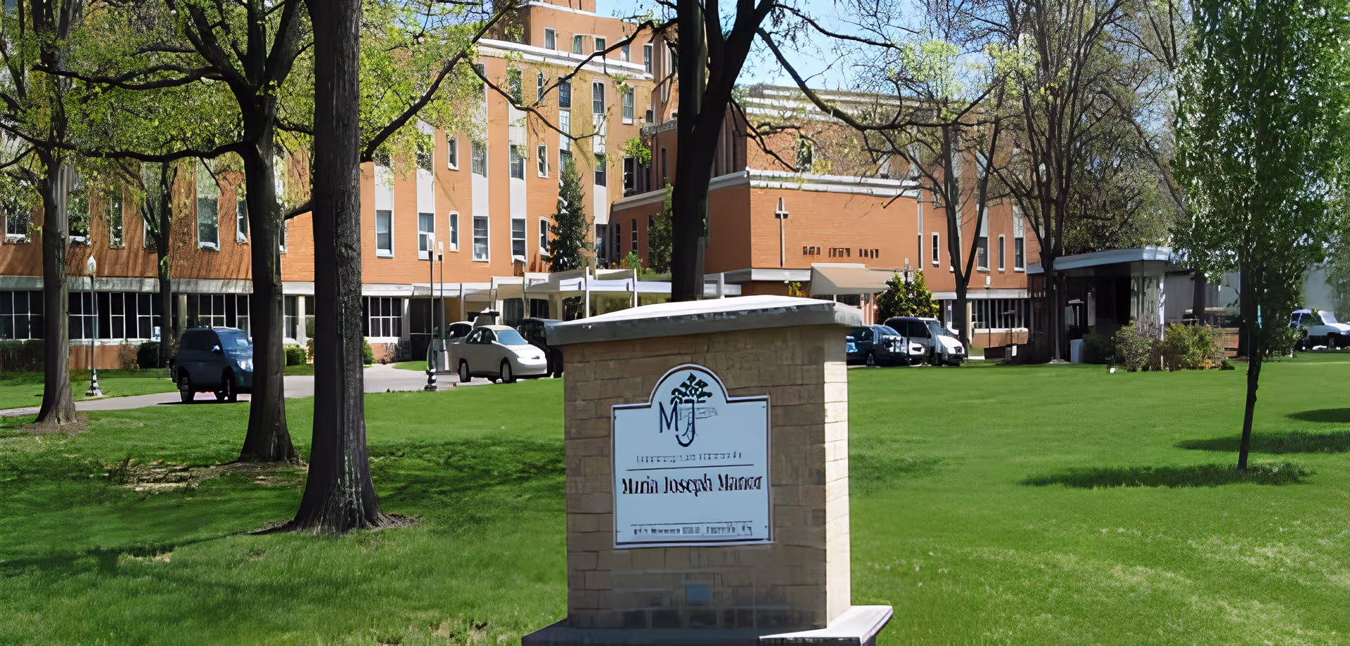 Exterior view of Maria Joseph Continuing Care Community building with a green lawn and several trees in the foreground. A sign with the facility's name is prominently displayed on a brick pedestal near the center of the image. Several cars are parked near the building under a clear sky.