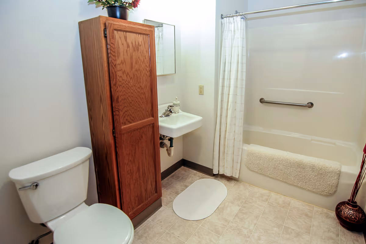 A clean bathroom featuring a white toilet, a wooden cabinet, a small white sink with a mirror above it, and a bathtub with a white shower curtain and a grab bar. A beige bath mat is placed on the floor in front of the sink, and a towel hangs over the edge of the bathtub.
