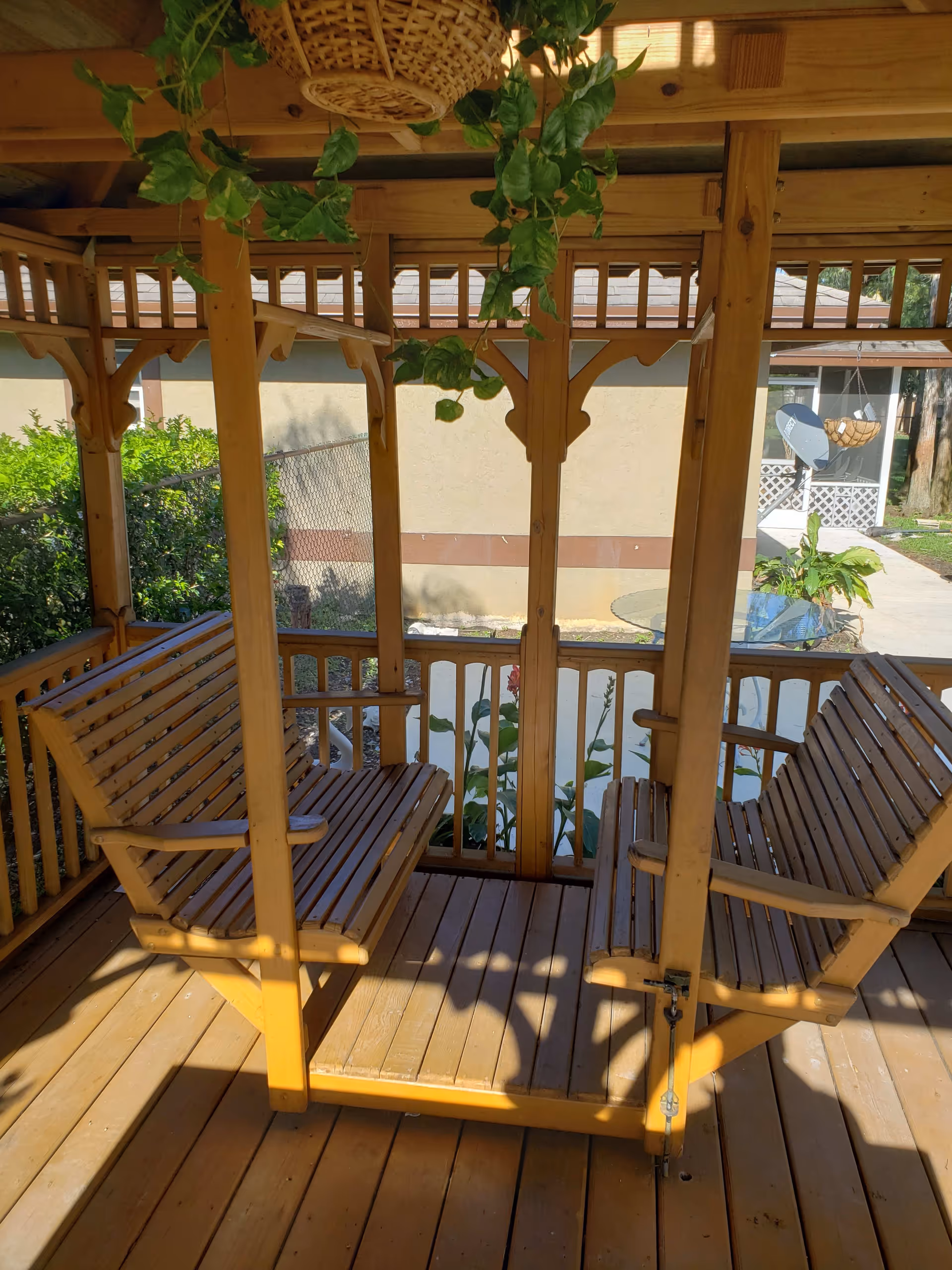 Wooden gazebo with two wooden bench swings facing each other, hanging plants above, and a view of a fenced garden and building exterior in the background.