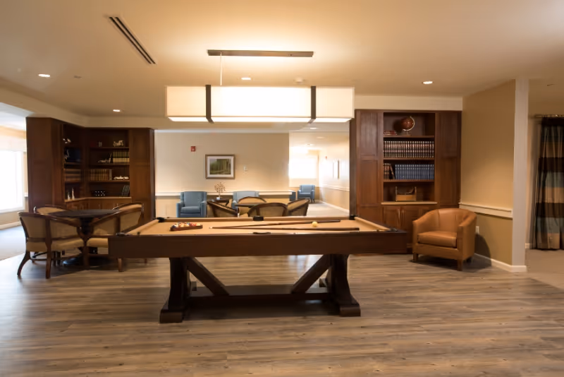 A spacious common area with a pool table in the center, surrounded by wooden bookshelves filled with books and decorative items. There are several seating arrangements including a round table with chairs, a leather armchair, and blue upholstered chairs in the background. The room has wooden flooring and warm lighting from a rectangular ceiling light fixture.