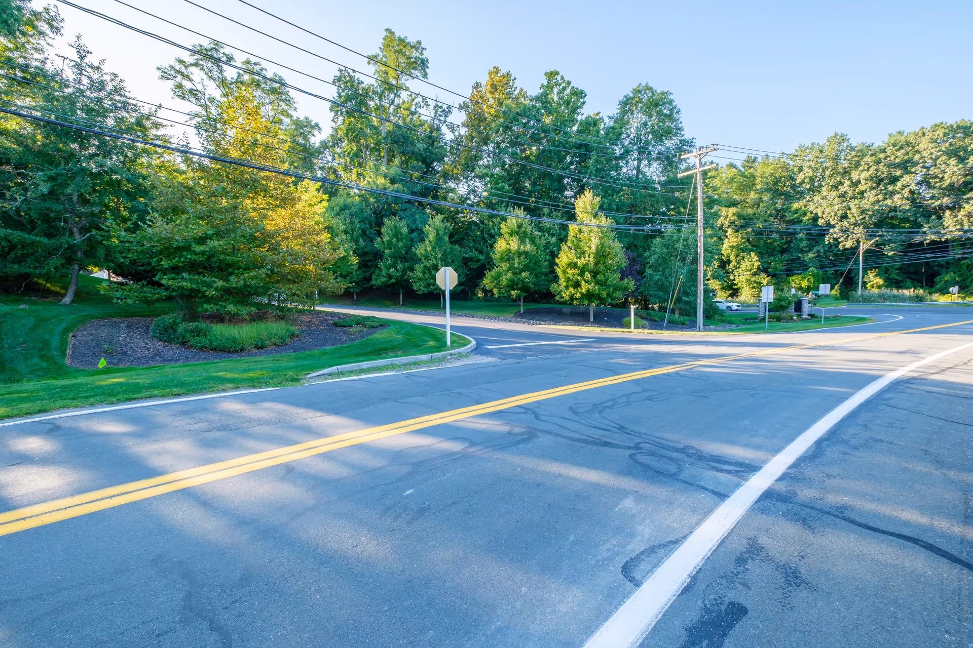 A paved road with yellow double lines and white edge lines curves through a green area with well-maintained grass and various trees under a clear blue sky. Utility poles and power lines run alongside the road.