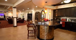 Interior view of a senior living facility featuring a kitchen area with dark cabinets, a curved island with bar stools, and a dining or lounge area with carpeted flooring and multiple seating arrangements in the background.