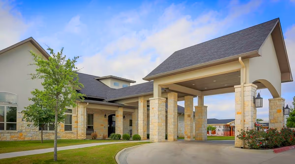 Exterior view of Arbor House Assisted Living and Memory Care facility showing a covered entrance supported by stone pillars, a driveway, green lawn, small trees, and a partly cloudy sky.
