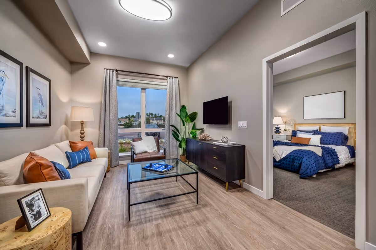 Modern furnished living room with a sofa, coffee table, TV console, and a view into a bedroom.