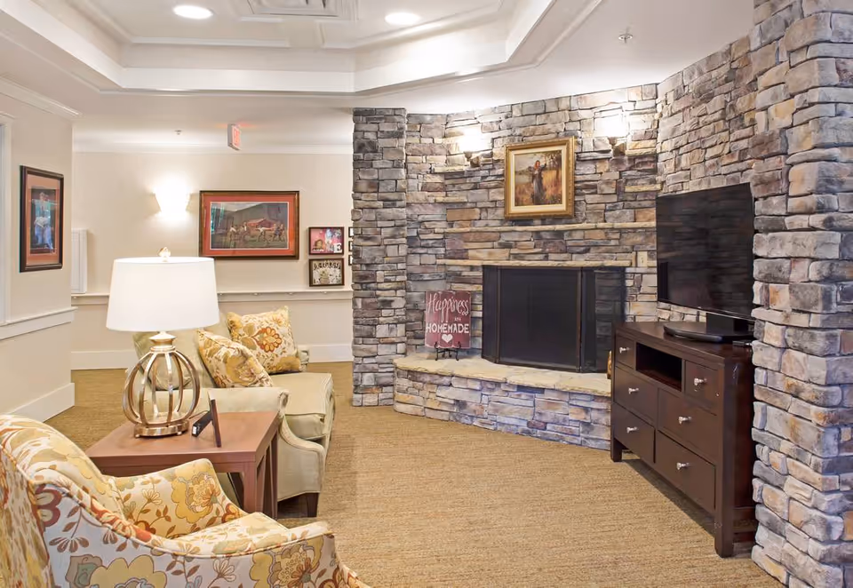 A cozy living room area with a stone fireplace and a flat-screen TV on a dark wooden stand. The room features beige carpet, a beige armchair with floral cushions, a side table with a lamp, and framed artwork on the walls. The fireplace has a decorative sign that reads 'Happiness is Homemade.'