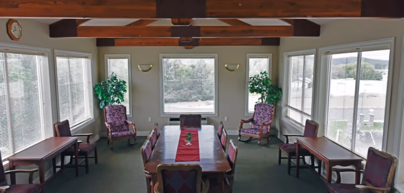 Sunlit common dining room with a long wooden table surrounded by chairs and armchairs, large windows, and exposed ceiling beams.
