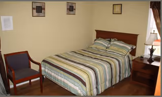 A bedroom with a double bed featuring a striped bedspread in shades of green, blue, and beige. There is a wooden nightstand with a lamp and a small decorative item next to the bed. A wooden chair with a dark cushion is positioned against the wall. The walls are painted light yellow and have three framed pictures hanging. A window with brown curtains is partially visible.