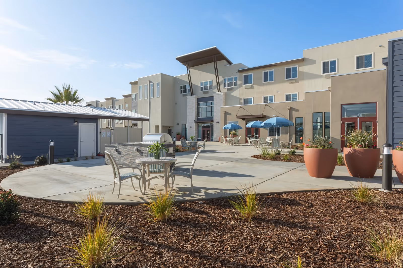 Outdoor patio area at WellQuest of Elk Grove featuring several tables with chairs and blue umbrellas, large potted plants, a built-in grill, and a modern multi-story building in the background under a clear blue sky.