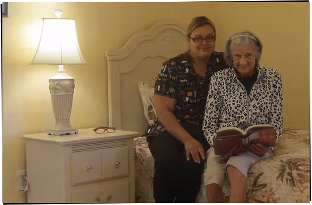 An elderly woman sitting on a bed reading a book with a caregiver sitting beside her in a warmly lit bedroom. A nightstand with a lamp and a pair of glasses is visible next to the bed.