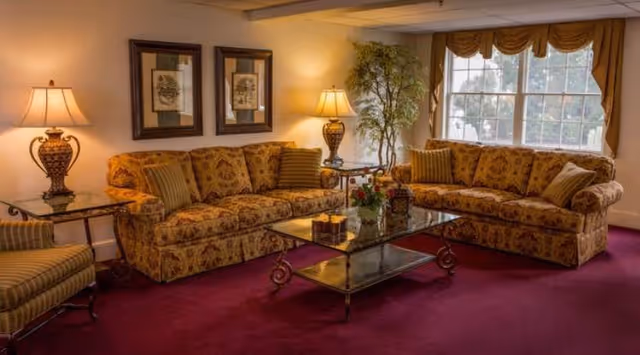 A cozy living room with two patterned sofas arranged in an L-shape around a glass coffee table. The room has a deep red carpet, two table lamps on side tables, framed artwork on the wall, a large window with gold curtains, and a tall potted plant in the corner.