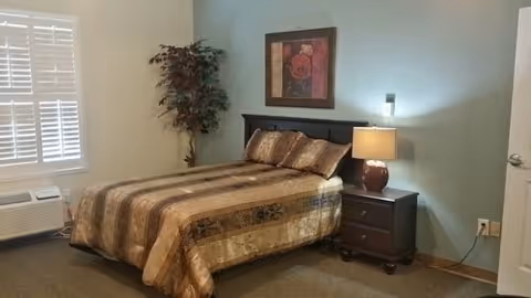 A bedroom with a bed covered in a brown and beige striped comforter, two matching pillows, a dark wooden headboard, a nightstand with a lamp, a framed floral picture on the wall, a potted plant in the corner, a window with white shutters, and a white air conditioning unit below the window.