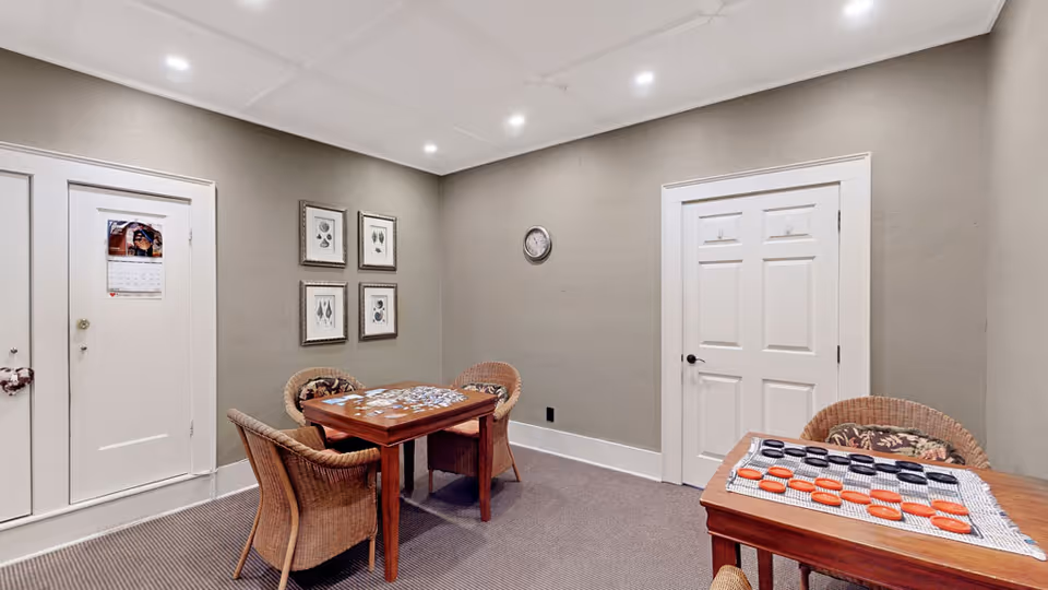 A small room with two wooden tables, each set up for a board game with checkers pieces. There are four wicker chairs around the tables. The walls are painted gray and decorated with four framed pictures and a clock. Two white doors are visible, and the floor is carpeted.