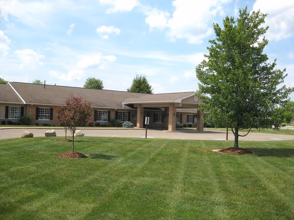 Exterior view of a single-story assisted living facility building with a covered entrance, surrounded by a well-maintained lawn with two small trees and a few large rocks. The sky is partly cloudy.