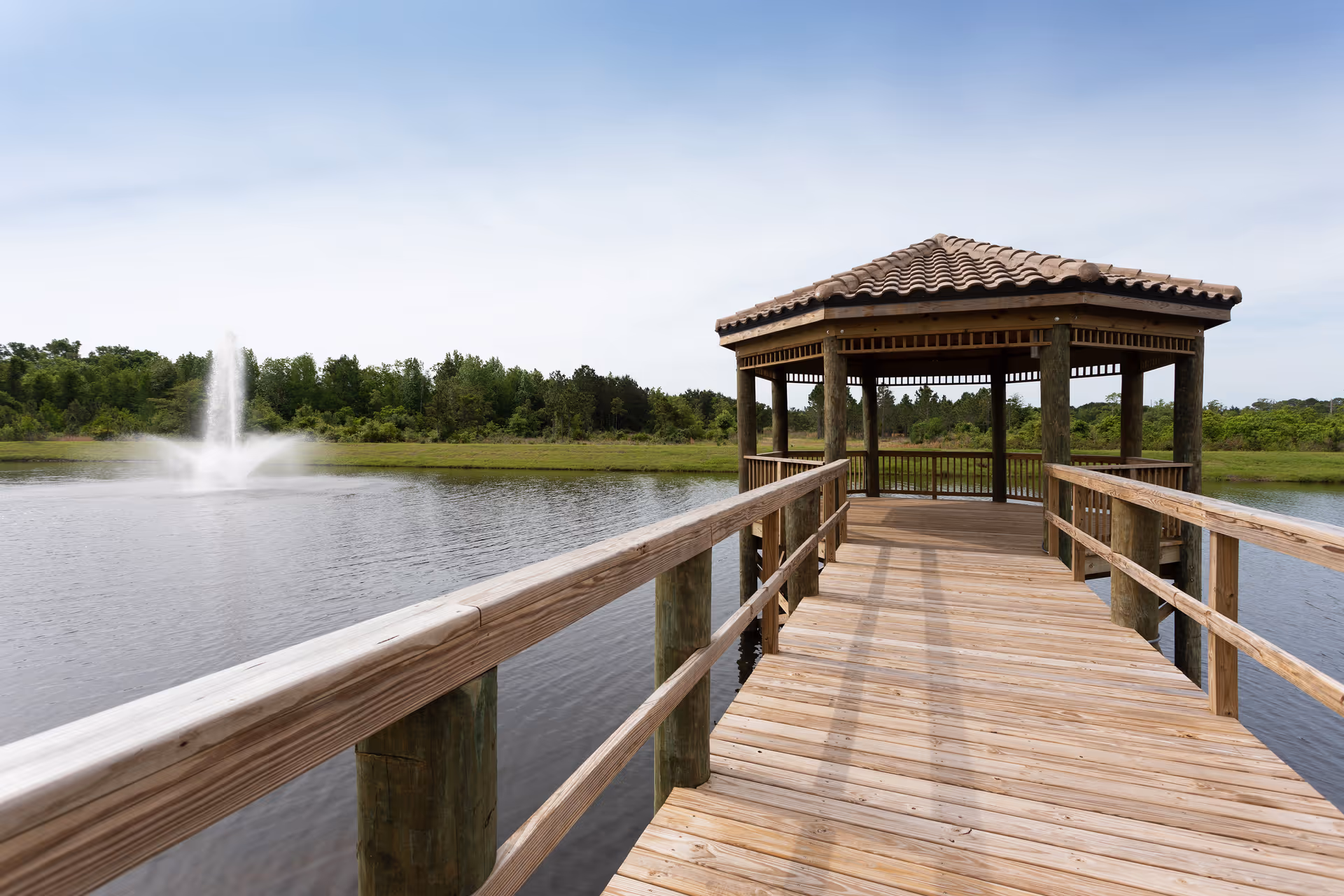 A wooden pier leading to a gazebo with a tiled roof over a calm pond with a water fountain in the background, surrounded by greenery and trees under a clear sky.