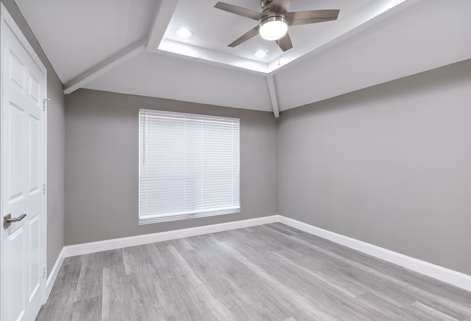Empty room with light gray walls, a window with closed blinds, a white door, gray wood flooring, and a ceiling fan with light in a recessed ceiling.
