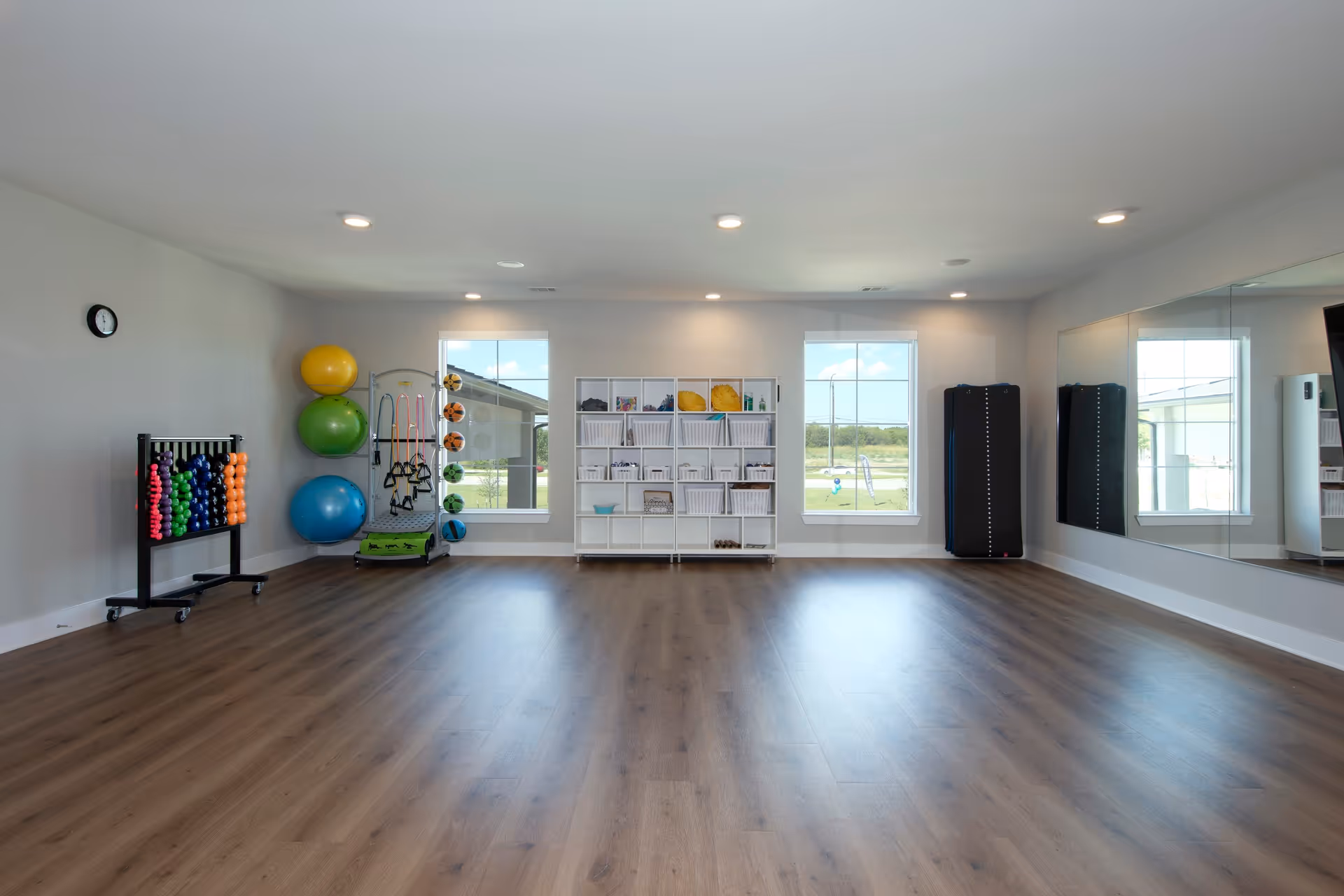 A spacious exercise room with wooden flooring, two large windows, and a wall mirror. The room contains various fitness equipment including colorful exercise balls, resistance bands, small weights, and storage shelves with baskets and other workout accessories. A clock is mounted on the wall.