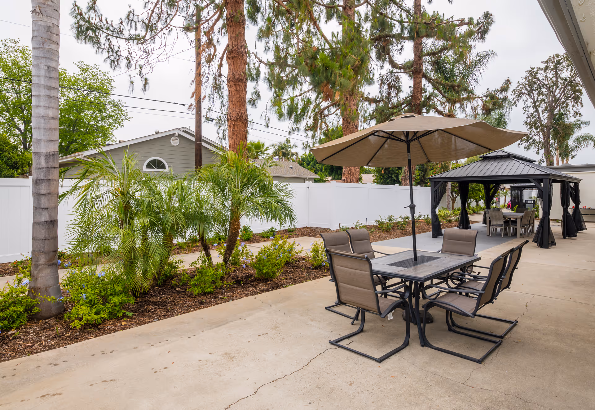 Outdoor patio area with a table and four chairs under a large umbrella, surrounded by trees and plants. In the background, there is a gazebo with another table and chairs underneath. The area is paved and enclosed by a white fence.