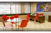 A bright and modern interior space with a glass round table surrounded by red chairs. In the background, there is a wooden desk with a computer and more red chairs. Large windows let in natural light, and a decorative plant and wall art featuring flowers add to the welcoming atmosphere.