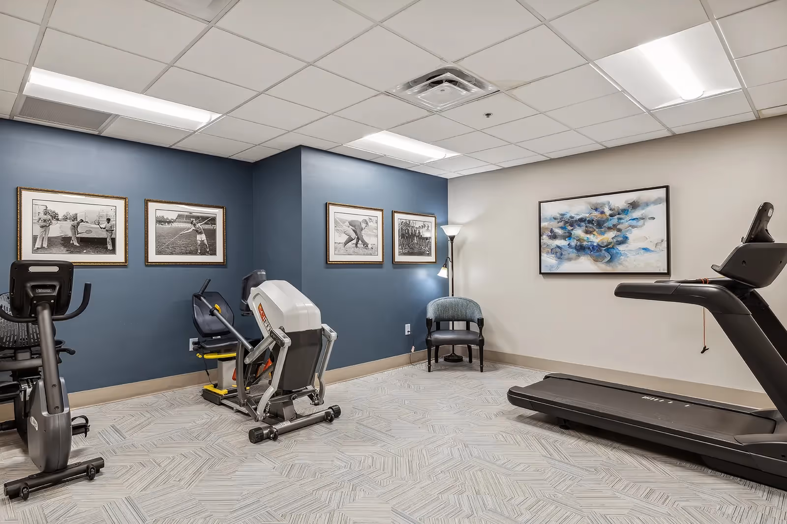 A fitness room with exercise equipment including a recumbent bike, a seated leg press machine, and a treadmill. The walls are painted blue and beige, decorated with framed black-and-white sports photographs and a colorful abstract painting. There is a single chair and a floor lamp in the corner.