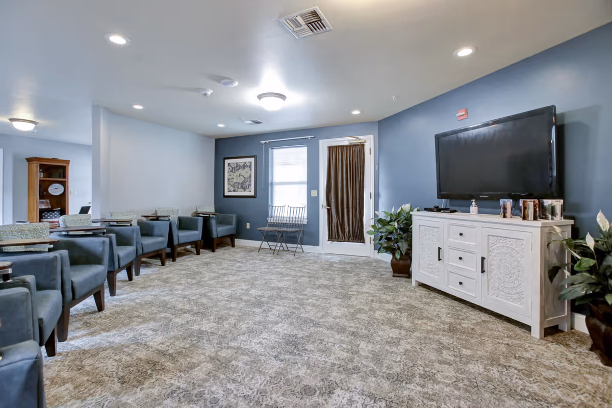 Bright senior living common room with rows of blue armchairs facing a wall-mounted TV and white console.