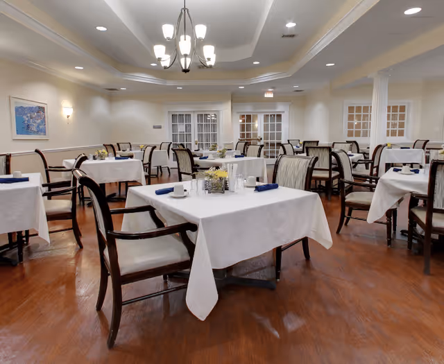 A dining room with multiple tables covered in white tablecloths, each set with cups, napkins, and small flower arrangements. The room has wooden floors, cushioned chairs, soft lighting from ceiling fixtures, and white walls with framed artwork.