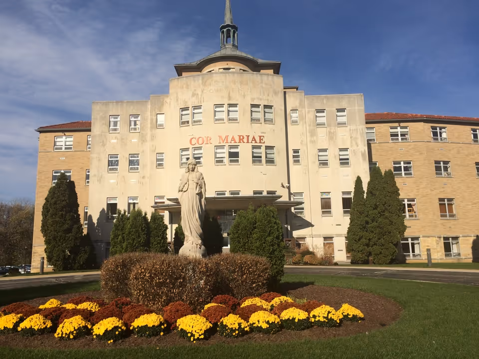 Front exterior view of a multi-story building with the words 'COR MARIAE' displayed prominently on the facade. In front of the building is a statue of a robed figure surrounded by neatly trimmed bushes and colorful flowers. The sky is clear and blue.