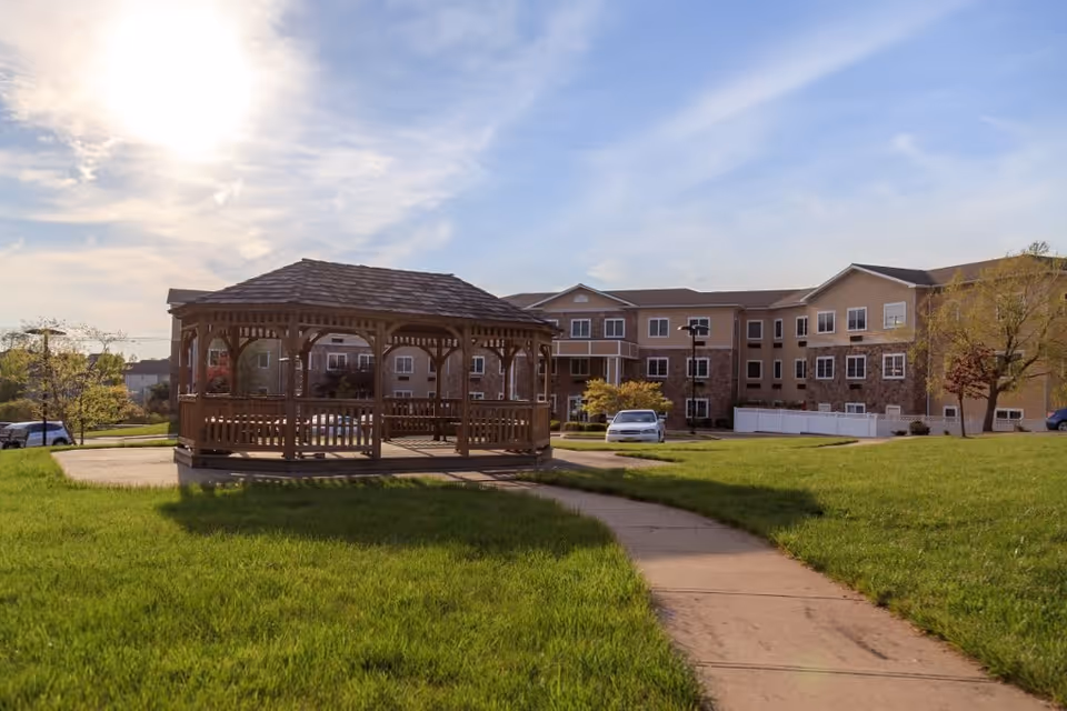A wooden gazebo situated on a concrete pad in a grassy outdoor area with a paved walkway leading to it. In the background, there is a multi-story residential building under a partly cloudy sky with the sun shining brightly.