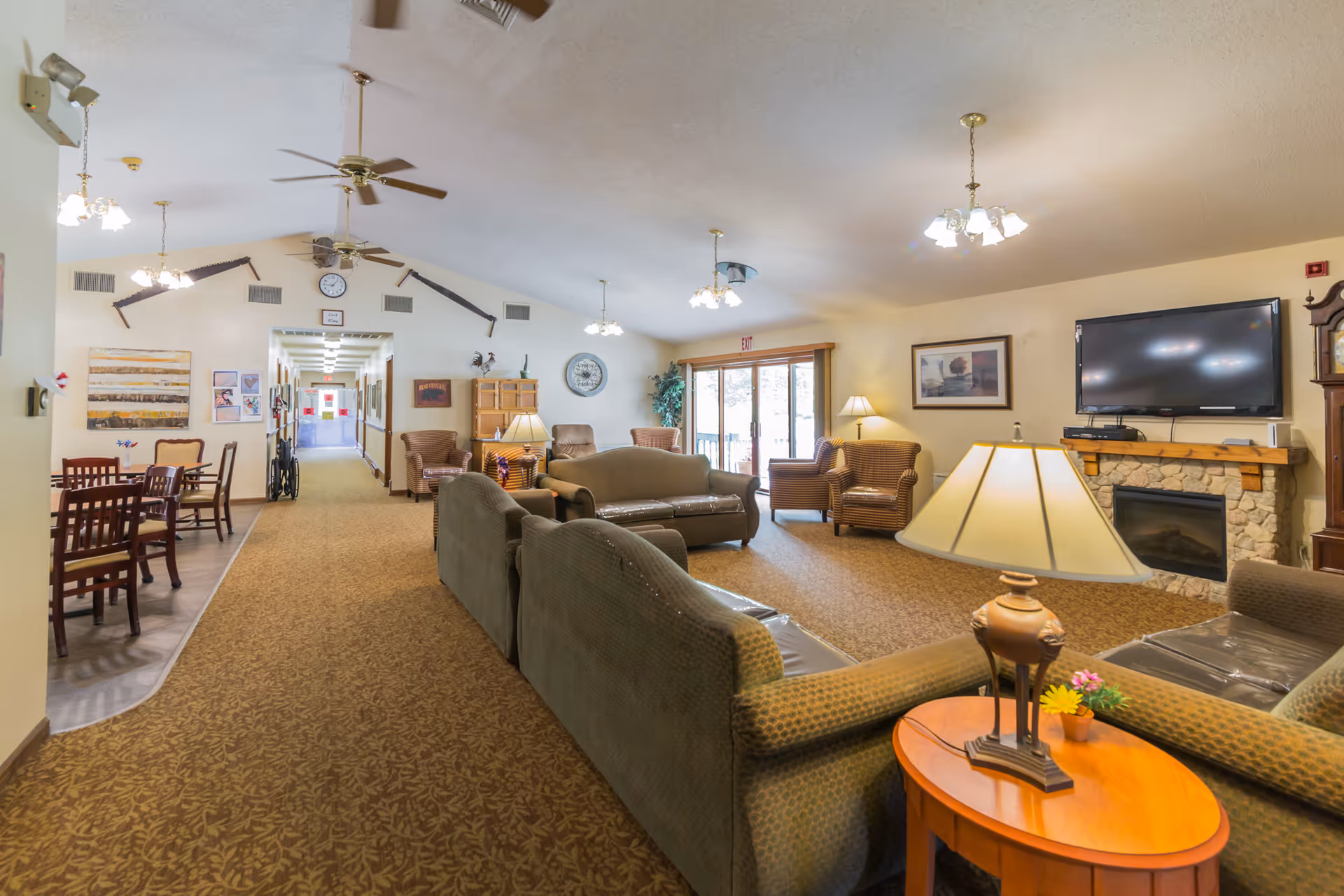 A spacious senior living facility common area with multiple green upholstered sofas and armchairs arranged around a stone fireplace with a mounted flat-screen TV. The room has carpeted floors, ceiling fans, and several hanging light fixtures. There is a hallway leading to other rooms, a dining area with tables and chairs on the left, and large sliding glass doors letting in natural light on the right.
