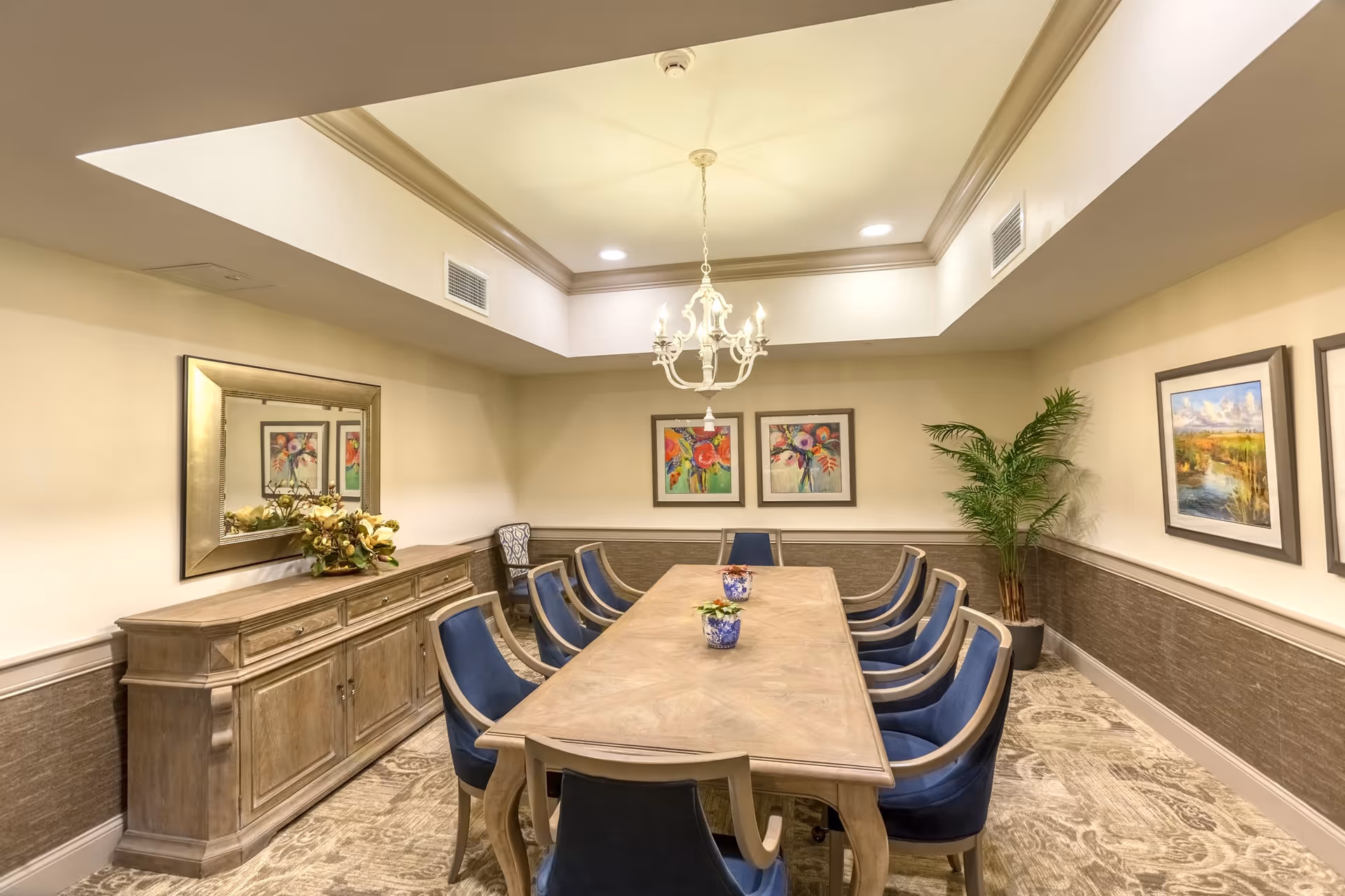 A well-lit dining room with a long wooden table surrounded by eight blue cushioned chairs. The room features a chandelier hanging from a recessed ceiling, a sideboard with a floral arrangement, framed colorful artwork on the walls, a large mirror, and a potted plant in the corner.