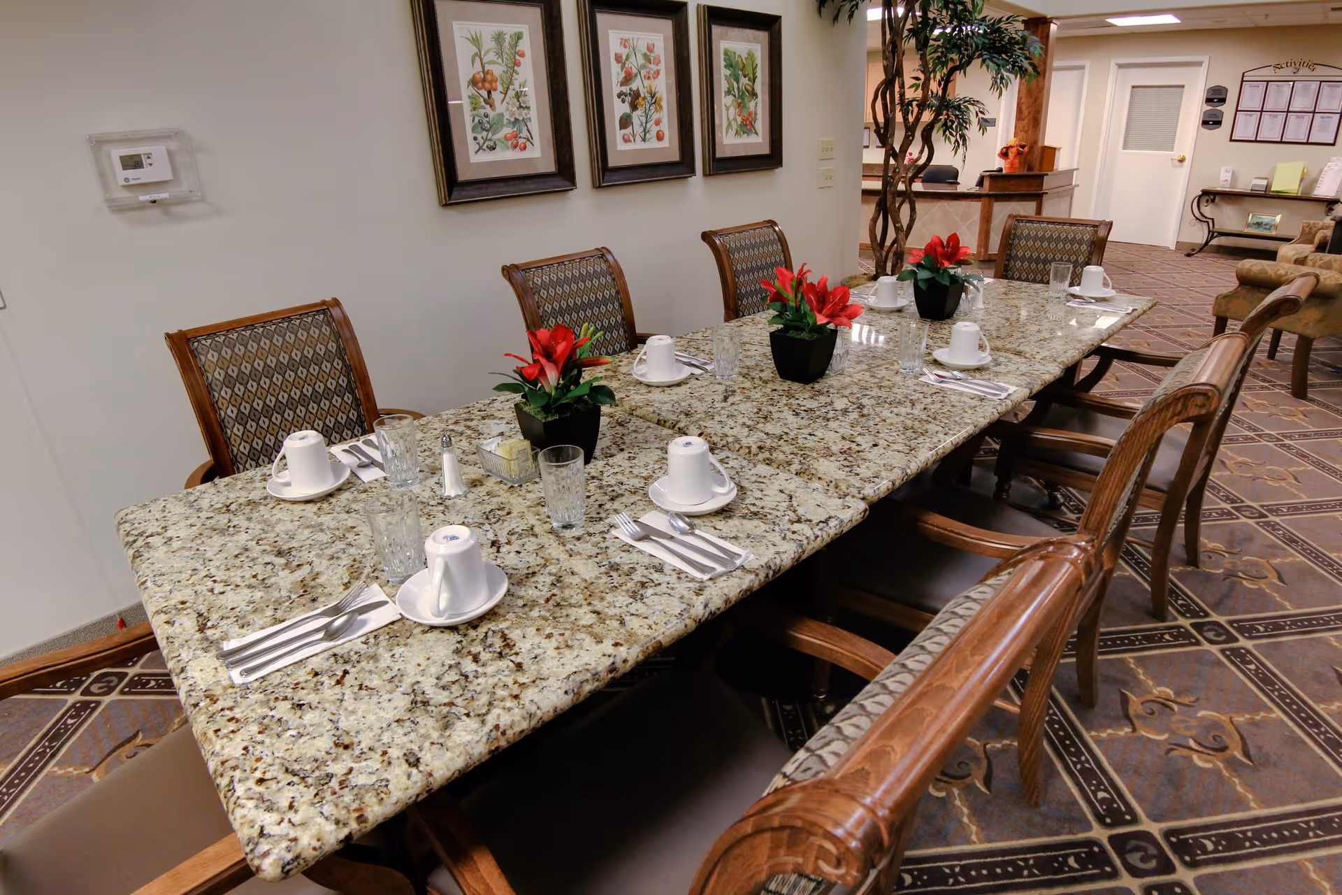 A long granite dining table set with white cups, glasses, silverware, and small potted red flowers in a well-lit room with patterned carpet and framed botanical prints on the wall.