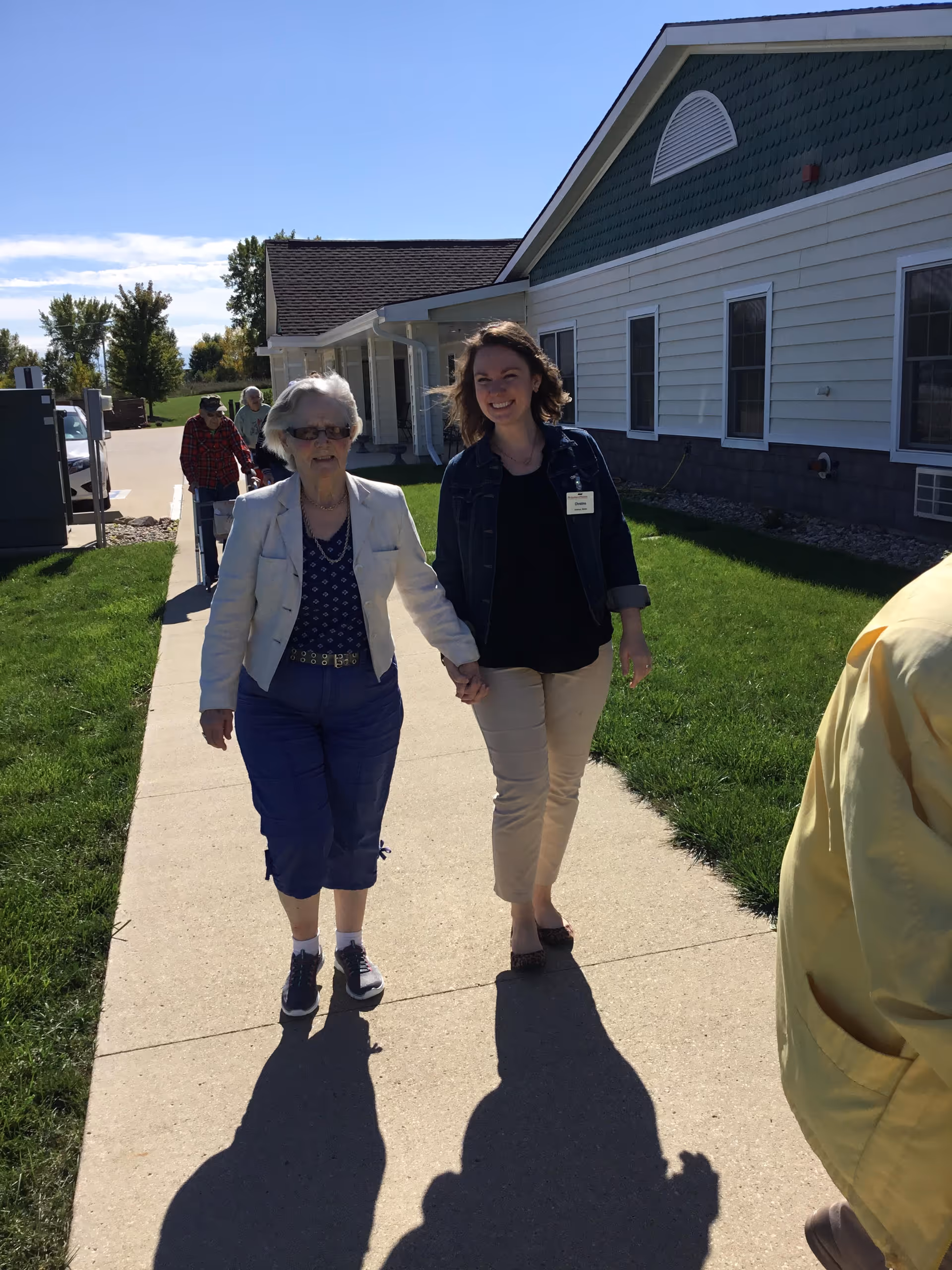 An elderly woman and a younger woman holding hands and walking on a sidewalk outside a single-story building with white siding and green trim on a sunny day. Other people are walking behind them, and there is green grass on either side of the sidewalk.