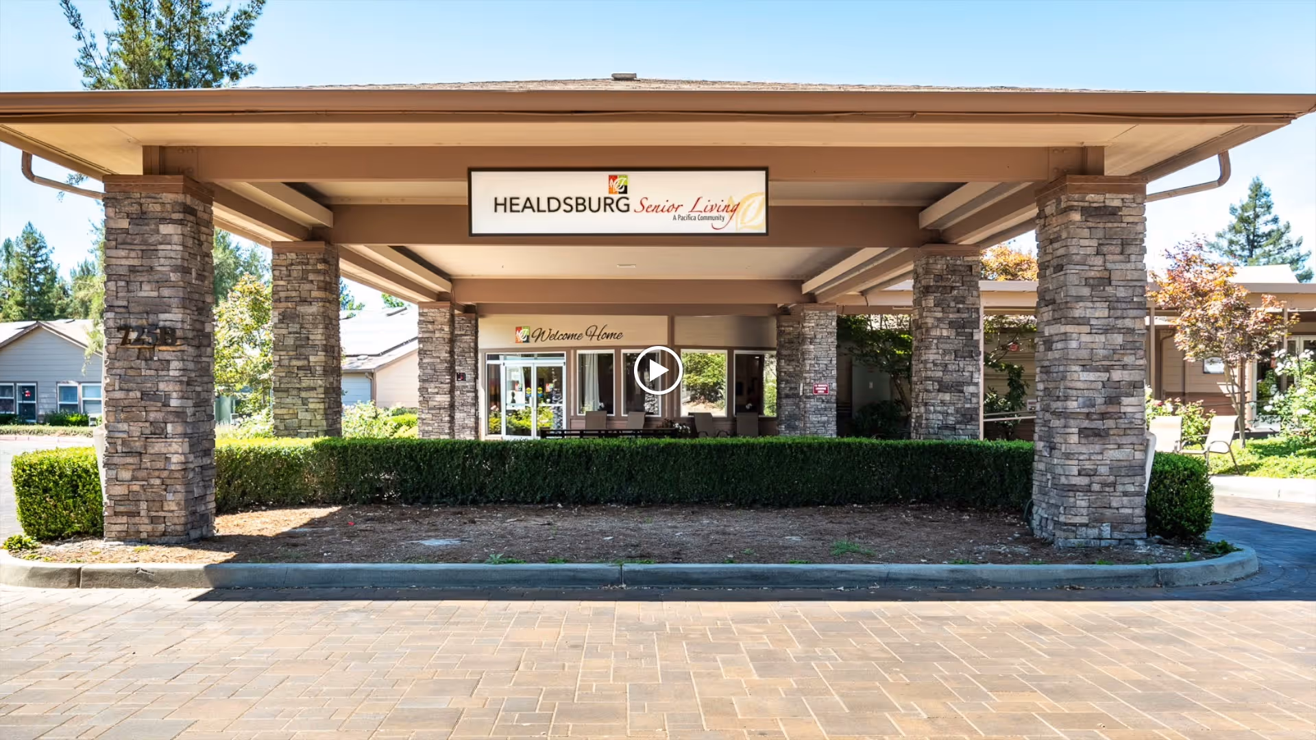 Entrance of Healdsburg Senior Living facility featuring a covered driveway supported by stone pillars, with a sign above reading 'Healdsburg Senior Living'. There are trimmed hedges and a paved driveway in front, and the building is surrounded by trees and other greenery.