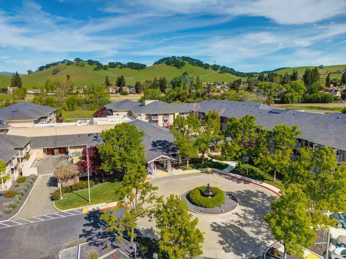 Aerial view of The Village at Rancho Solano senior living facility showing multiple connected buildings with gray roofs surrounded by green trees and landscaped grounds. A circular driveway with a central fountain is visible in front of the main entrance. Rolling green hills and a blue sky with scattered clouds are in the background.