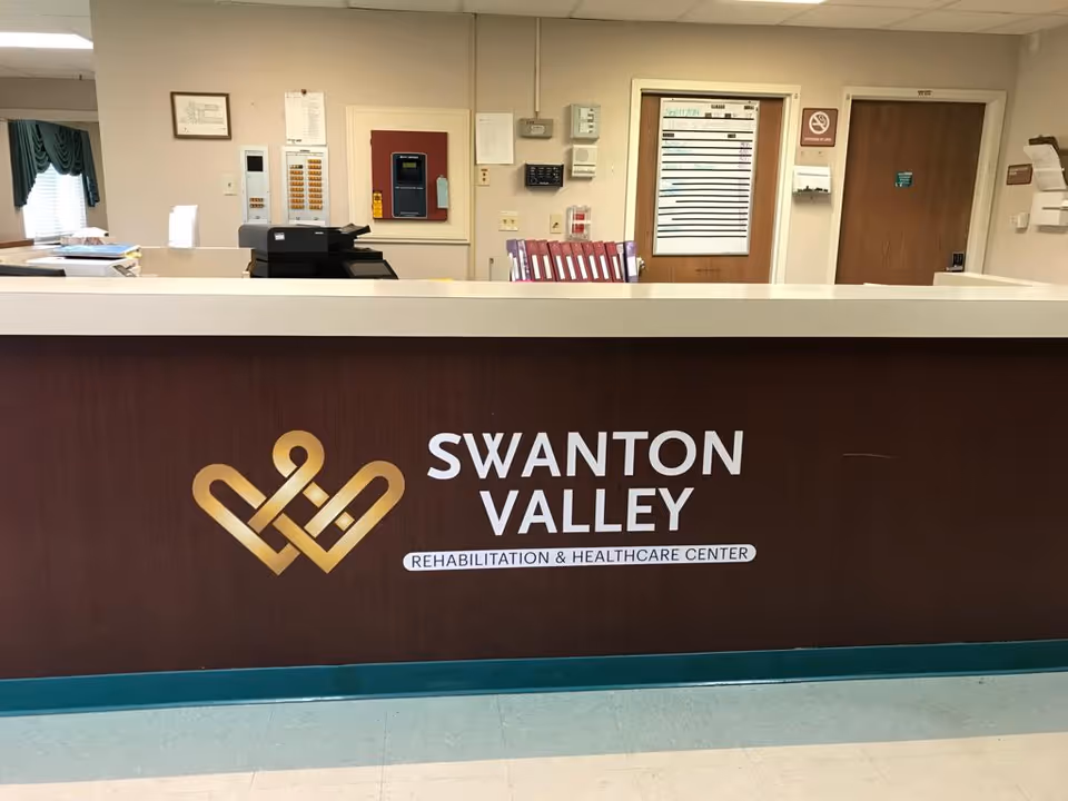 Reception desk area inside Swanton Valley Rehabilitation and Healthcare Center with a brown front panel displaying the facility's name and logo. Behind the desk are office equipment, a bulletin board, and doors.